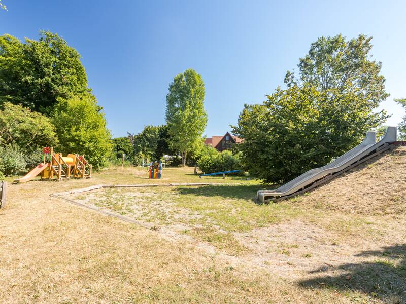 Außenspielbereich auf Wiese mit Wellenrutsche rechts im Bild, Kletterturm auf der linken Seite und der Wippe im Hintergrund. Rundherum sind Bäume.