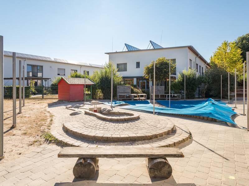 Große gepflasterte Matschstraße mit Sandkiste im Hintergrund. Vorne ist eine Sitzbank zu sehen im Hintergrund zwei Gebäude und Bäume.