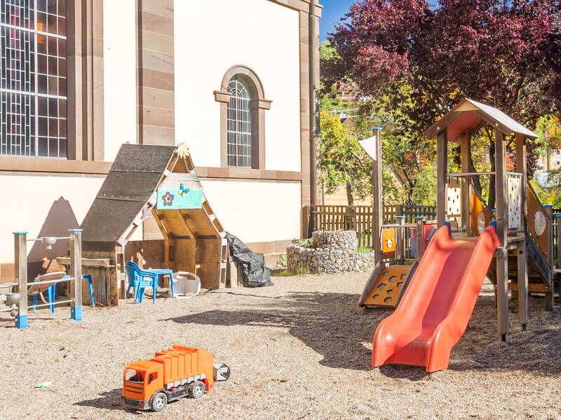 Ein kleiner Kletterturm mit Rutsche und eine Matschstraße auf sandigem Untergrund. Ein kleiner orangener Spiel-LKW  und eine Reckstange sind zu sehen. Hinter dem Zaun stehen ein paar Bäume.