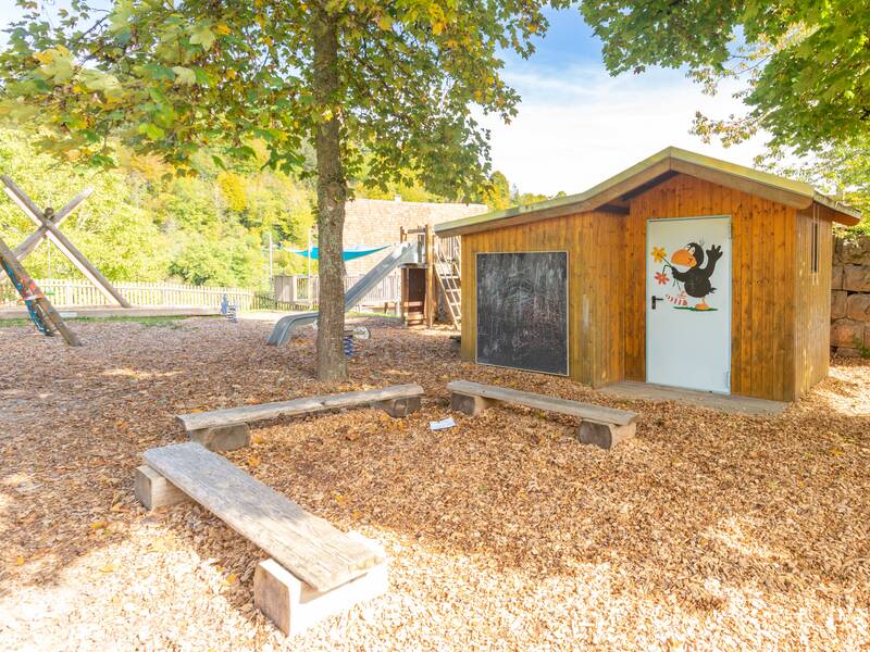 Drei Sitzbänke in U-Form vor der Hütte für Spielgeräte. Im Hintergrund steht die Rutsche, Schaukel und ein Klettergerüst. Bäume rundherum.