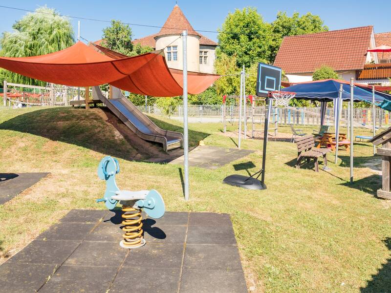 Zwei Wipptiere auf Fallschutzmatten sowie ein Schaukeltier im Vordergrund. Eine Rutsche die an einem begrasten Hang runterführt. Eine kleine Holzhütte sowie einige Sitzgelegenheiten in einem Pavillon und unter einem Sonnensegel. Im Hintergrund stehen mehrere Bäume und Gebäude.