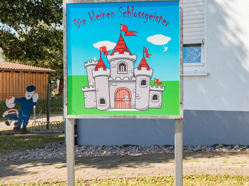 Zum Eingang des Kindergartens weißt ein Schild mit der Aufschrift "Die kleinen Schloßgeister".  Im Hintergrund am Zaun ist aus Holz ein Polizist mit erhobener Hand und ein kleiner Junge angebracht.