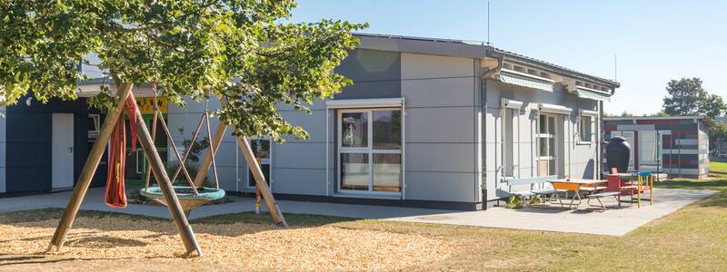 Vor einem grauen Gebäude mit großen Fenstern steht ein Nestschaukel auf einer Wiese unter einem Baum. An der rechten Gebäudeseite ist eine betonierte Terrasse auf der ein paar Sitzbänke und Tische platziert sind. Ein weiteres Gebäude steht im Hintergrund.