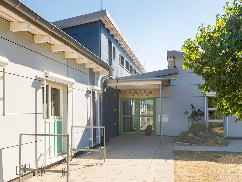 Der Eingangsbereich des Kindergartens ist gepflastert. An der Hauswand steht ein Fahrradständer. Der Eingang ist überdacht. In diesem Bereich steht ein Blumenkübel. Rechts an der Hauswand steht ein Busch. Es ist eine Rasenfläche und ein größerer Baum zu erkennen. 