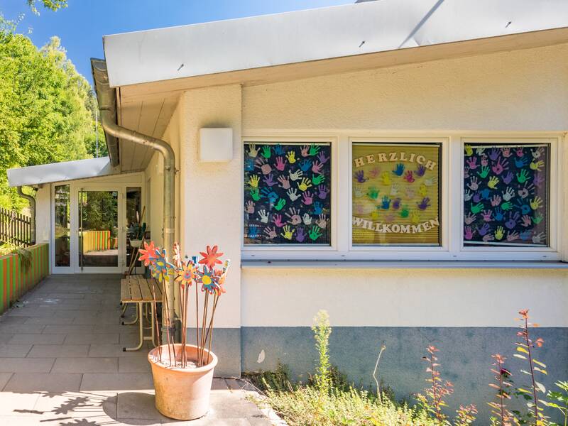Der Eingangsbereich des Kindergartens ist teilweise überdacht und führt am Gebäude vorbei. Vorne sind drei große Fenster, die mit Kinderhänden bemalt sind. An der Ecke steht ein Blumenkübel mit bunten Holzblumensteckern. Links ist eine bunte Mauer und viele Bäume zu sehen.
