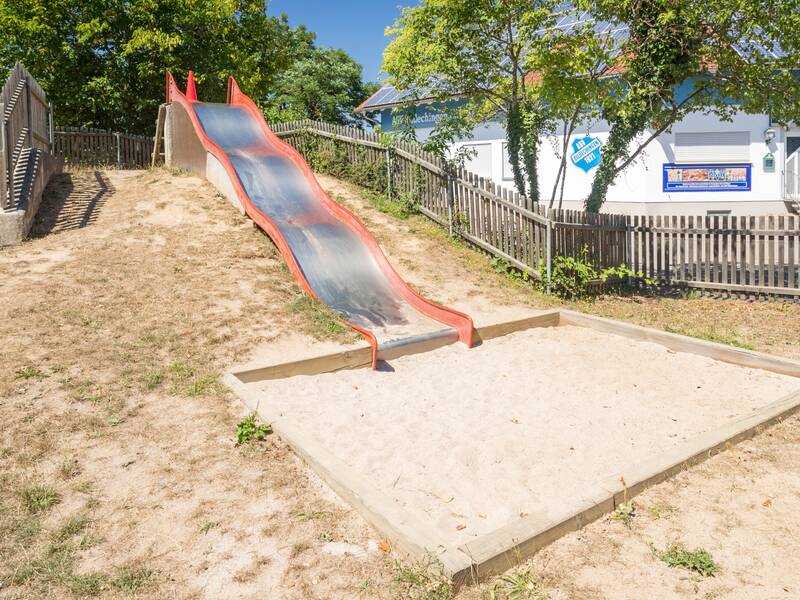 Große Wellenrutsche aus Metall die von einem Hang runter führt und in einer Sandkiste endet. Das Gelände ist eingezäunt. Es sind einige Bäume und ein Gebäude zu sehen.