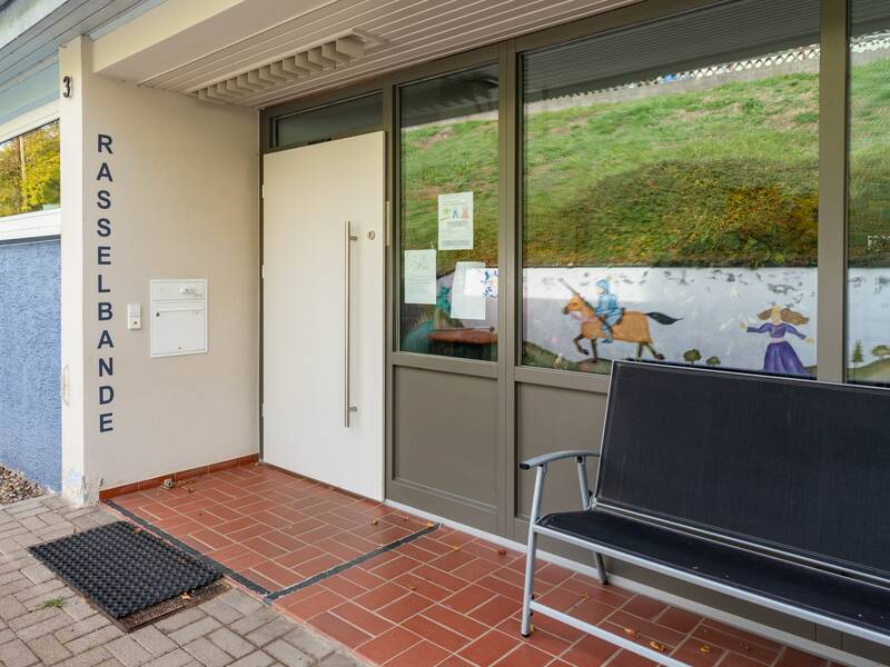 Der Eingangsbereich des Kindergartens ist überdacht. Der Boden davor ist gepflastert, der überdachte Teil gefliest. Die Fliesen sind ein bisschen erhöht. Neben der Eingangstür steht eine Sitzbank. Die Fenster sind mit Kinderbildern geschmückt. 