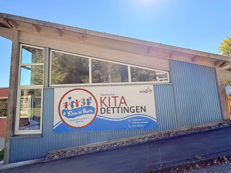 Das Bild zeigt ein modernes Gebäude mit blauer Verkleidung, großen Fenstern und einem Schild mit der Aufschrift "Städtische KITA Dettingen".