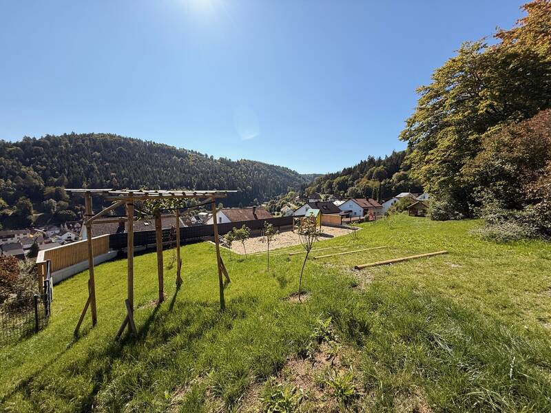 Eine grüne Wiese mit einem Holzgerüst im Vordergrund bietet einen Ausblick auf ein Dorf, umgeben von bewaldeten Hügeln unter einem klaren blauen Himmel.
