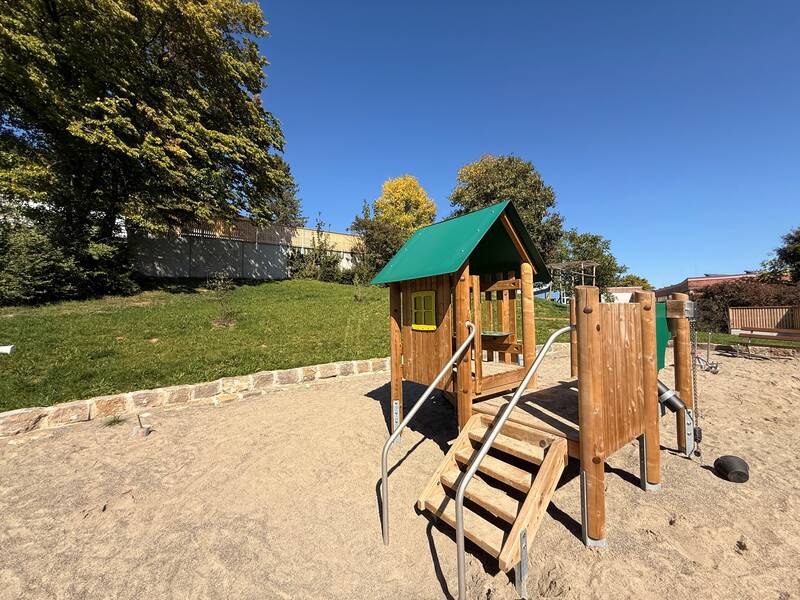 Auf dem Bild ist ein kleiner Spielplatz mit einer Holzspielstruktur und Rutsche zu sehen, umgeben von Sand und Bäumen im Hintergrund.
