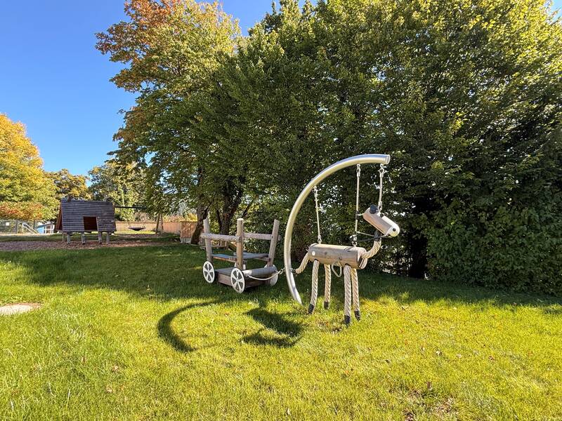 Ein Spielplatz mit einer hölzernen Schaukel in Form eines Pferdes und einem hölzernen Wagen auf einer Wiese, umgeben von grünen Bäumen.