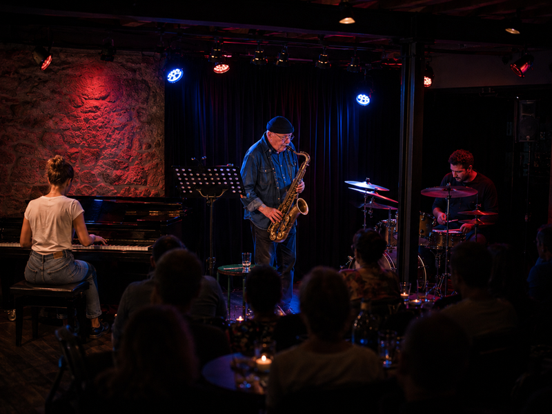 In einem gemütlichen, dunklen Club spielen drei Musiker auf einer kleinen Bühne: eine Person am Klavier, eine mit einem Saxophon und eine am Schlagzeug, während das Publikum zuschaut.