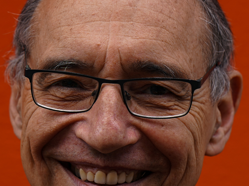 Ein lächelnder älterer Mann mit Brille vor einem orangefarbenen Hintergrund.