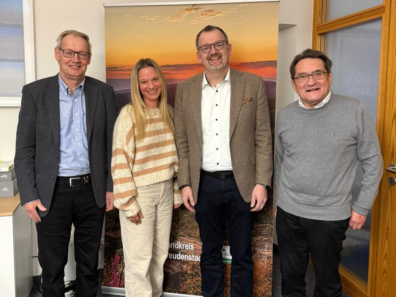 Vier Personen stehen nebeneinander vor einem Banner mit Landschaftsbild und Text, zwei Männer tragen Anzüge, eine Frau einen gestreiften Pullover, ein Mann einen grauen Pullover. Auf dem Bild von links: Konrad Fassnacht, Katrin Lohner, Landrat Andreas Junt, Andreas Bronner