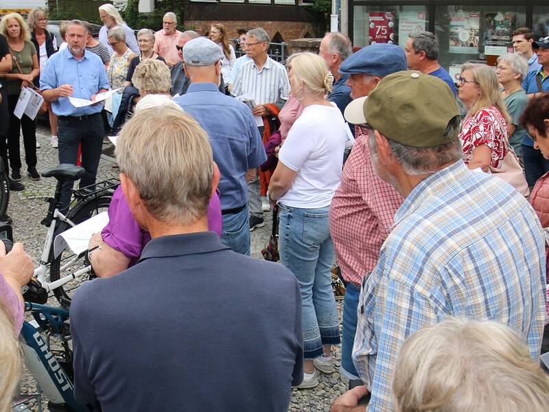 Eine Gruppe von Menschen steht im Freien zusammen, während eine Person in einem blauen Hemd zu ihnen spricht.