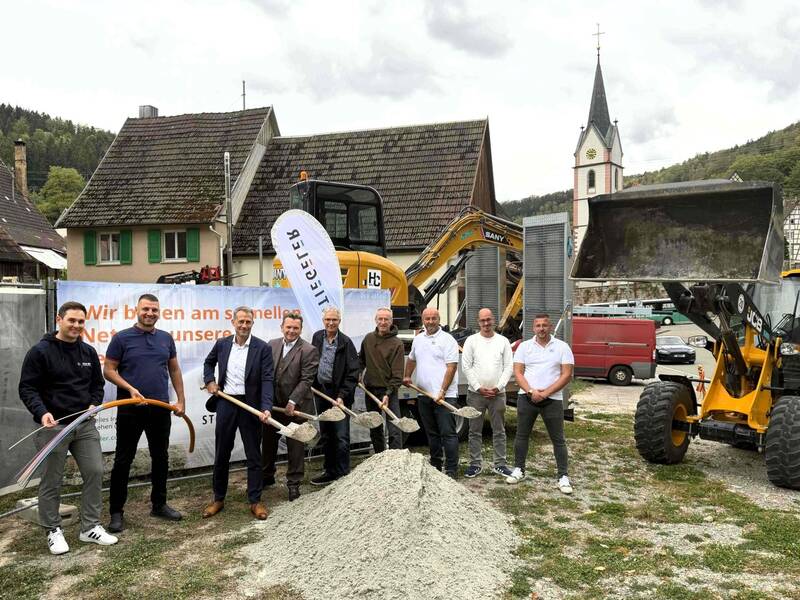 Eine Gruppe von acht Männern steht mit Schaufeln auf einem Baugelände vor Baggern und einem Banner, das auf ein Bauprojekt hinweist, im Hintergrund sind traditionelle Häuser und eine Kirche zu sehen.