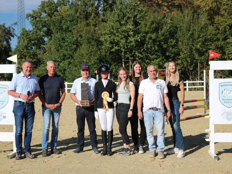 Eine Gruppe von acht Personen, darunter eine im Reitoutfit mit Medaille, posiert lächelnd auf einem Reitplatz zwischen Schildern des Reitervereins Nordstetten Horb e.V.
