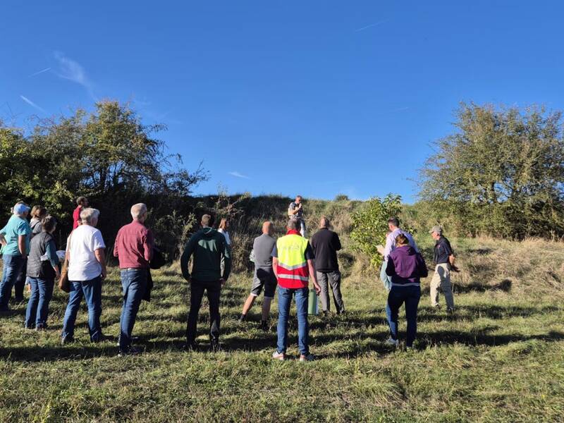 Eine Gruppe von Menschen steht auf einer Wiese vor einer erhöhten, mit Büschen bewachsenen Landschaft unter einem klaren blauen Himmel.
