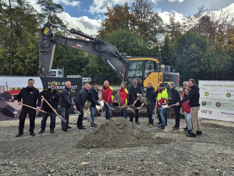 Eine Gruppe Menschen steht mit Schaufeln vor einem Bagger und einem kleinen Sandhaufen, während sie bei einer Grundsteinlegung auf einer Baustelle posieren.