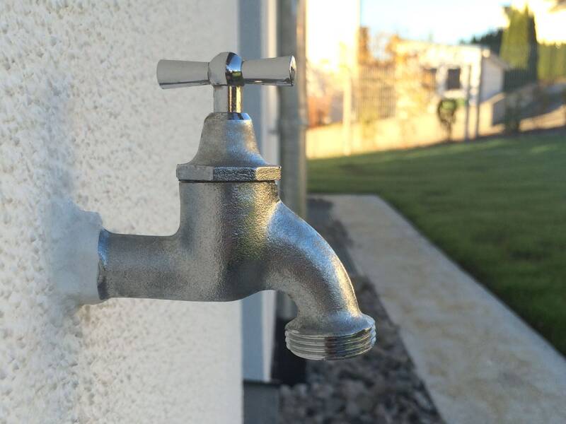 Ein silberner Wasserhahn ist an einer weißen Wand im Freien montiert, mit unscharfem Hintergrund einer Rasenfläche und Bäumen.