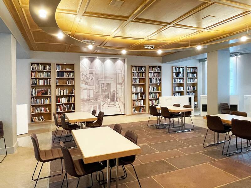 Ein Raum mit Stühlen und Tischen. Im Hintergrund Bücherregale und ein großes Gemälde.