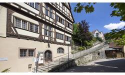 Eine alte Fachwerkfassade mit Erkern steht an einer gepflasterten Straße neben einer steinernen Treppe, die zu einem erhöhten Gehweg mit Bäumen und anderen Gebäuden führt.