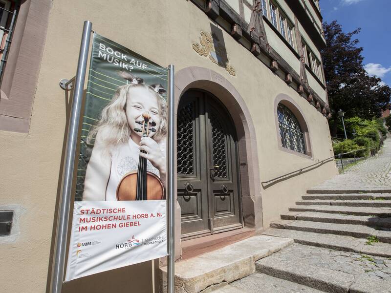 Ein Plakat vor einem historischen Gebäude wirbt für die Städtische Musikschule Horb am Neckar und zeigt ein lächelndes Kind mit einer Geige.