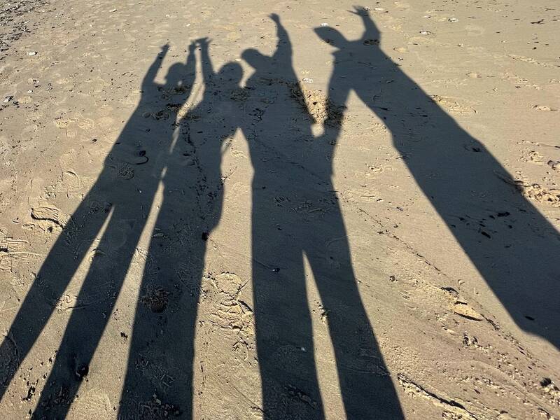 Schatten von vier Personen auf Sand, die Arme in verschiedenen Positionen heben.