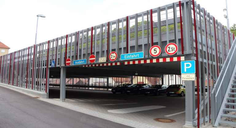 Das Bild zeigt ein mehrstöckiges Parkhaus mit Schildern für Einfahrt, Ausfahrt und Höhenbegrenzungen sowie geparkten Autos im unteren Bereich.