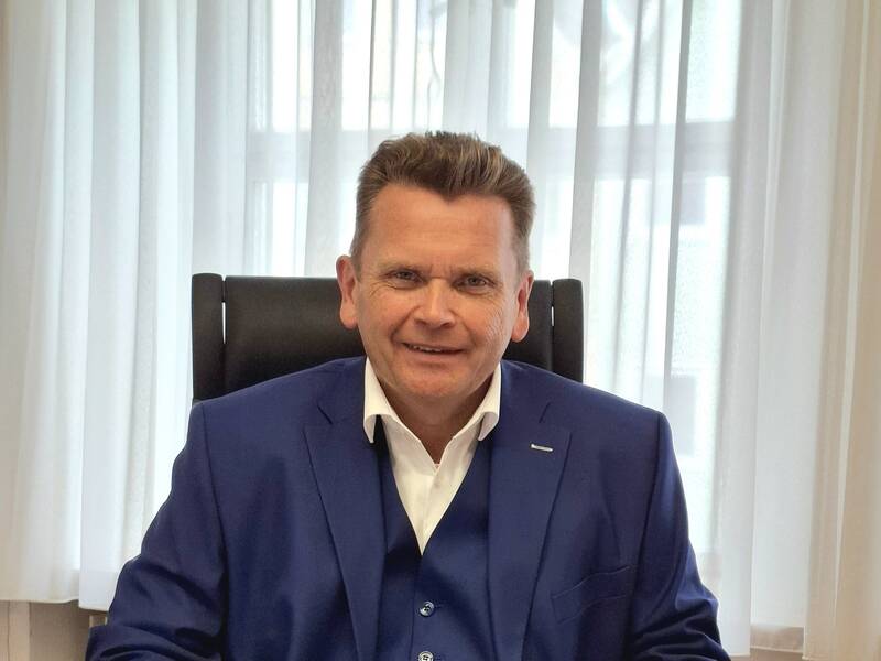 Mann in blauem Anzug sitzt lächelnd an Schreibtisch mit Dokument vor sich, helle Vorhänge im Hintergrund.