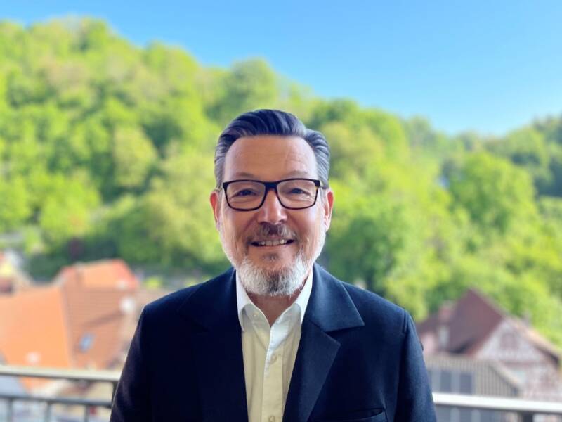 Mann mit Brille und Bart lächelt vor einem Balkon mit Blick auf grüne Bäume und Häuser im Hintergrund.