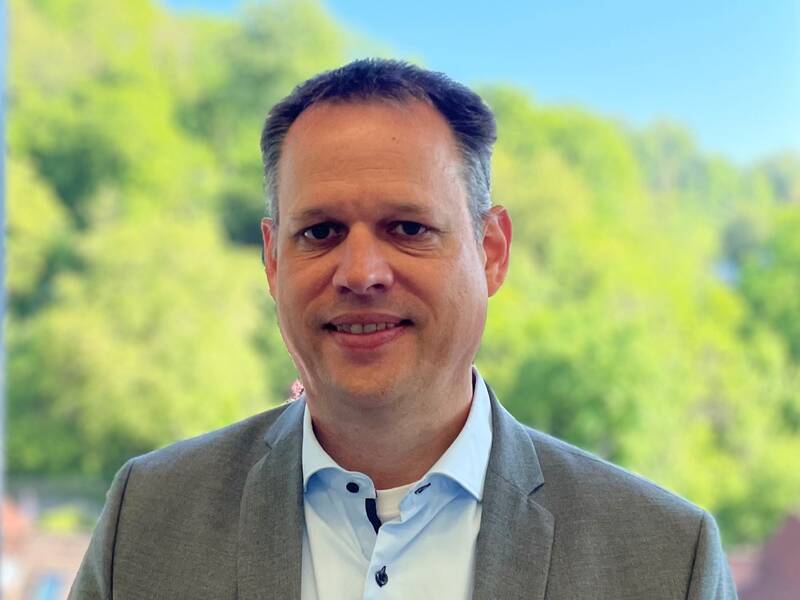 Mann in grauem Sakko und weißem Hemd steht vor großen Fenstern mit Blick auf grüne Bäume und Häuser.