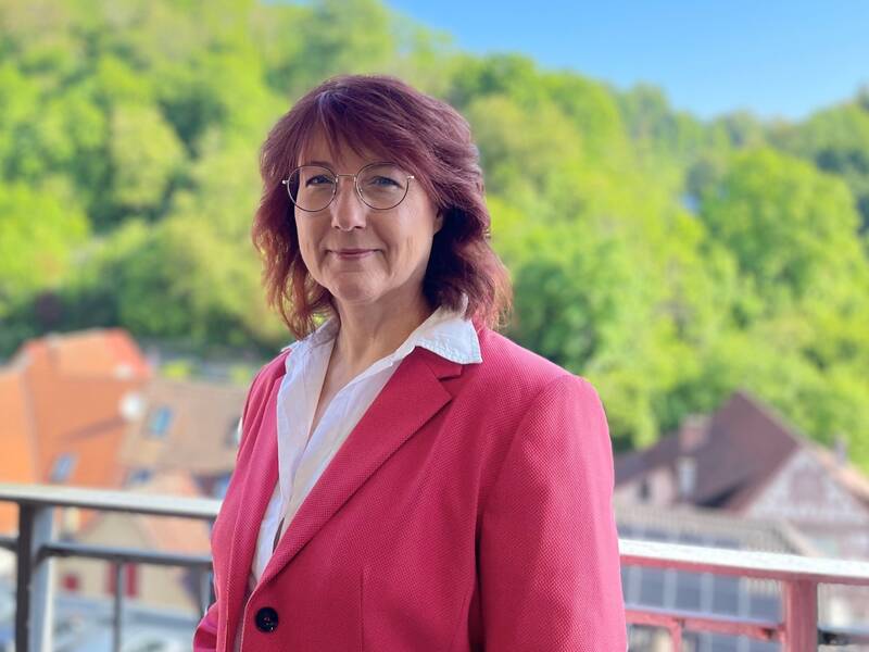 Frau mit roten Haaren und Brille trägt roten Blazer und weiße Bluse, steht auf Balkon mit Blick auf grüne Bäume und Häuser.