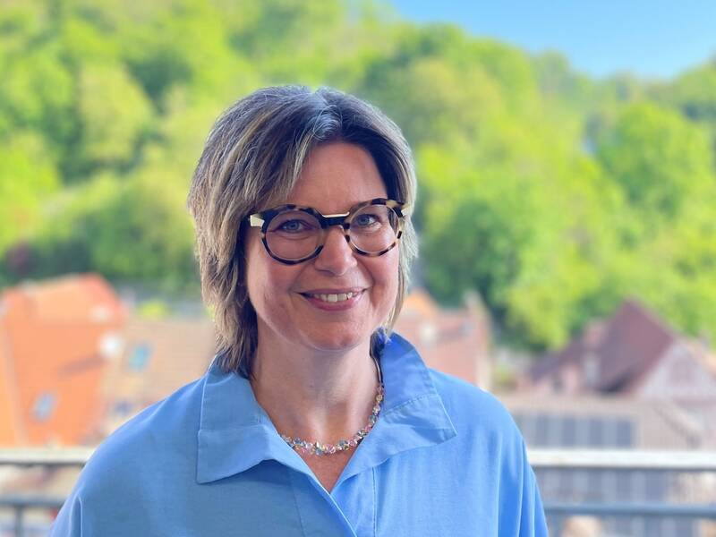 Frau mit Brille und blauer Bluse lächelt auf Balkon mit grünem Wald und Häusern im Hintergrund.
