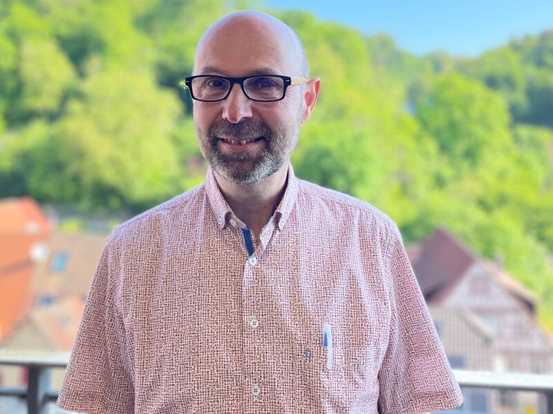Mann mit Brille und Hemd steht auf Balkon mit Blick auf grüne Hügel und Häuser im Hintergrund.