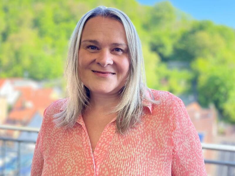 Frau mit langen blonden Haaren und rosa gemustertem Hemd lächelt vor einem Balkon mit grünem Wald im Hintergrund.