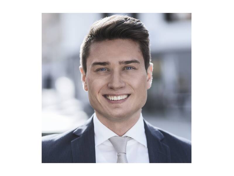 Ein lächelnder Mann in einem Anzug steht draußen, der Hintergrund ist unscharf.