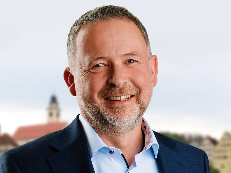 Ein Mann mit grauem Haar und Bart lächelt in die Kamera, gekleidet in ein blaues Sakko und ein helles Hemd, vor einem verschwommenen Hintergrund.