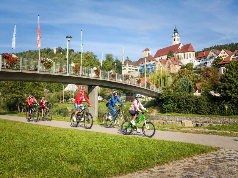 Eine Gruppe von Radfahrern fährt einen Weg entlang, während im Hintergrund eine Brücke mit Blumenschmuck und eine Kirche der Stadt Horb am Neckar zu sehen sind.