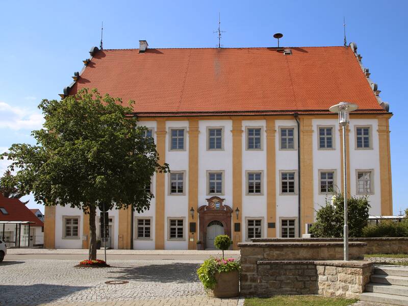 Auf dem Bild ist ein historisches Gebäude mit rotem Ziegeldach auf einem Marktplatz zu sehen.  Vor dem Gebäude steht ein Baum.