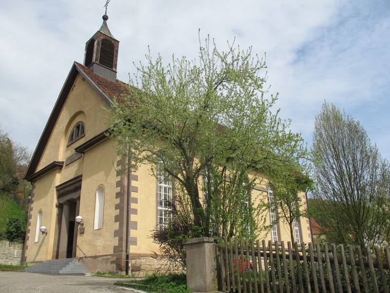 Eine hellgelbe Kirche umgeben von Bäumen und Sträuchern. Rechts im Bild sieht man einen braunen Gartenzaun.