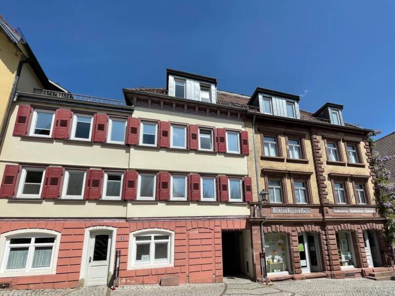 Zwei nenebeinander stehende, historische Gebäude mit roten Fensterläden unter blauem Himmel.