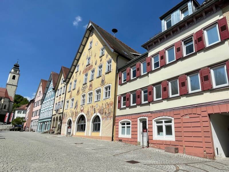 Mehrere historische Gebäude an einander gereiht mit einer Kirche im Hindergrund und blauem Himmel.