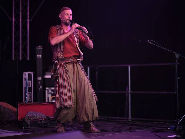Ein Mann in einem Bühnenoutfit spielt ein Blasinstrument auf einer dunklen Bühne.