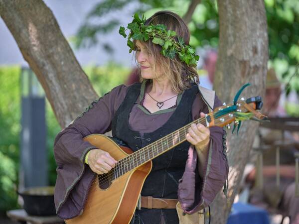 Eine Person mit Blumenkranz auf dem Kopf spielt Gitarre in einem gewandähnlichen Kostüm im Freien.