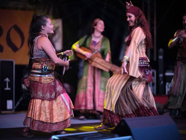 Eine Gruppe von Menschen in farbenfrohen, traditionellen Kostümen tanzt und spielt Musikinstrumente auf einer Bühne.