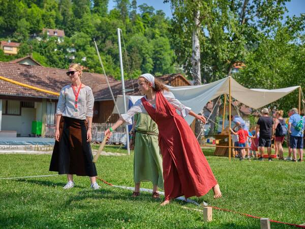 Eine Frau in einem mittelalterlich anmutenden Kleid wirft draußen auf einer Wiese ein Holzstück, während eine andere Frau daneben steht und zusieht.