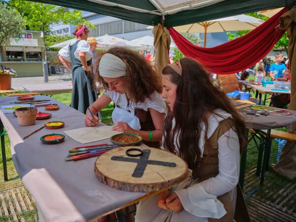 Zwei Mädchen basteln und zeichnen an einem Tisch im Freien, während sie historische Kleidung tragen und von Kunstmaterialien umgeben sind.