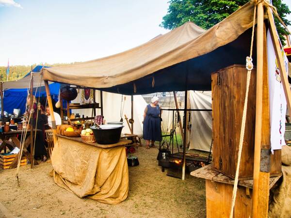Ein mittelalterlich wirkender Marktstand mit einem Zelt und Tischen, auf denen Obst und Geschirr stehen, während eine Person in historischer Kleidung daneben steht.