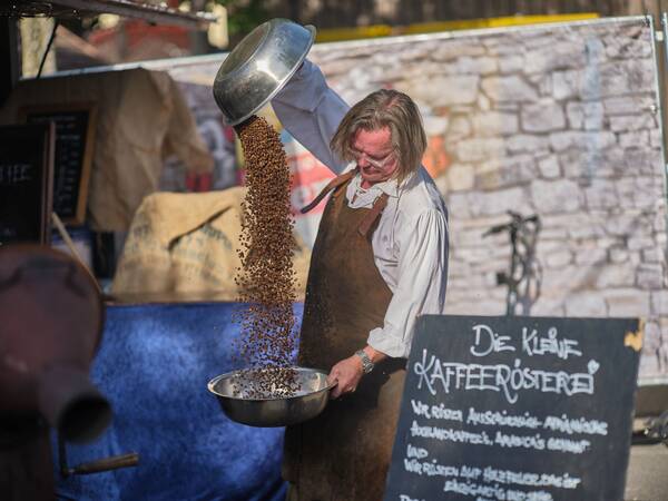 Ein Mann in einer Schürze gießt Kaffeebohnen aus einer großen Schüssel in eine kleinere Schüssel, während eine Tafel mit der Aufschrift "Die kleine Kaffeerösterei" zu sehen ist.
